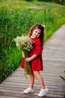 Kestane rengi elbiseli, küçük, uzun saçlı bir kız elinde papatya buketiyle ahşap bir köprüde duruyor. Bir buketle oynuyor. Seçici odak resmi