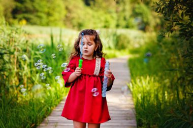 Üzerinde sabun köpükleri olan kırmızı elbiseli küçük siyah saçlı bir kız tahta bir köprüde duruyor ve sabun köpüğü üflüyor..