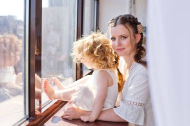 Anne ve kızı pencere eşiğinde pencereden dışarı bakıyor. Büyük bir pencerenin arkasında mutlu bir aile var..