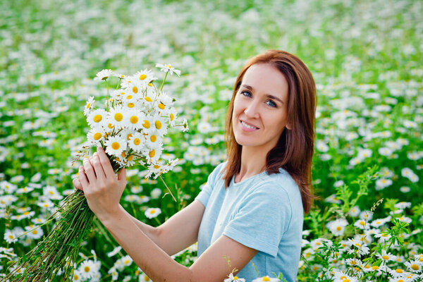 A young smiling brunette in a blue dress holds a bouquet of daisies in her hands and sits on a chamomile field. He looks at the camera. Copy space.