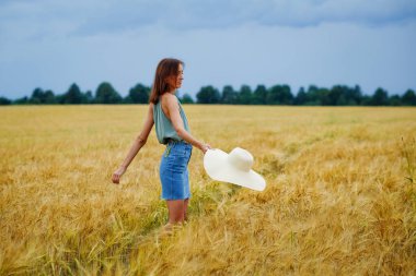 Elinde şapkası olan zayıf bir kız gökyüzünün arka planında bulutlarla buğday tarlasının ortasında dönüyor. Kırsal yaz manzarası.