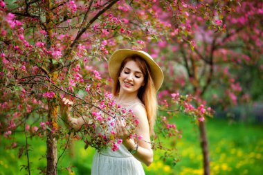 Güzel, genç, kızıl saçlı bir kadın, çiçek açan ağaçların arka planında baharda açtığı bahçenin güzel kokusunun tadını çıkarıyor. Gün batımında doğada gülümseyen genç bir bayan. Kırsal konseptte bahar