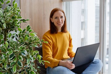 Sarı kazaklı bir çalışan ya da öğrenci elinde dizüstü bilgisayarla pencerenin kenarında oturuyor. Uzaktan çalışmak rahat ve elverişlidir..