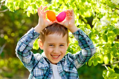 Çiçekli bir bahar bahçesinde Paskalya yumurtası avında bir çocuk. Süslü yumurtaları tavşan kulağı gibi kafasına dayar..