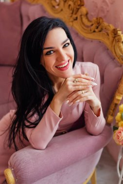 Pembe elbiseli, gülümseyen bir bahar kızının portresi. Uzun saçlı ve gülümseyen inanılmaz bir esmer elinde çiçeklerle kanepede oturuyor. Sevgililer Günü ve Kadınlar Günü