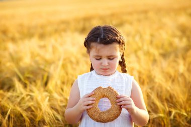 Saçları siyah örgülü küçük bir kız gün batımında buğday tarlasında simit yiyor.