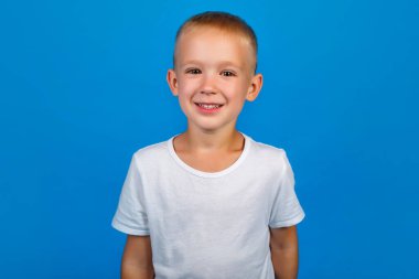 Beyaz tişörtlü mutlu bir çocuğun portresi. Arka planda mavi ile izole edilmiş kameraya bakıyor.