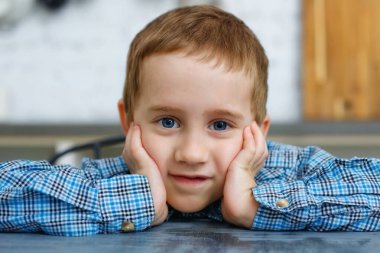 Düşünceli sarışın bir çocuk mutfak masasında oturur ve kameraya bakar. Yüzünde elleri olan bir çocuğun büyük bir portresi..