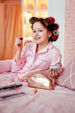 Mutlu çocuk. Pembe pijamalı güzel bir kız yatak odasındaki yatakta makyaj yapıyor. Kız kafasında bigudilerle ve ellerinde makyaj fırçalarıyla gülüyor.