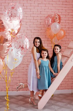 Gülümseyen bir anne ve iki kız kardeş benzer giysiler içinde pembe bir odada pembe şeftali balonları ve merdiven..