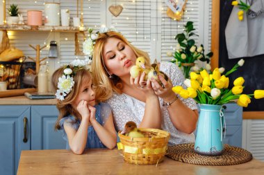 Paskalya kompozisyonu. Başlarında çiçek çelenkleri olan anne ve kız mutfak masasında oturup ördek yavrularıyla oynuyorlar. Bir kadın elinde bir ördek yavrusu tutuyor..