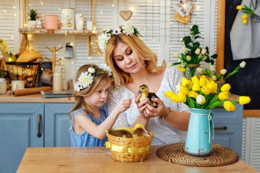 Paskalya kompozisyonu. Başlarında çiçek çelenkleri olan anne ve kız mutfak masasında oturup ördek yavrularıyla oynuyorlar. Bir kadın elinde bir ördek yavrusu tutuyor..