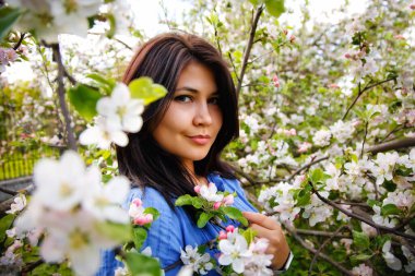 Bahar çiçekli bir bahçede mavi elbiseli, koyu renk saçlı, çekici, iri yarı bir kadının yakın plan portresi. Bahar.