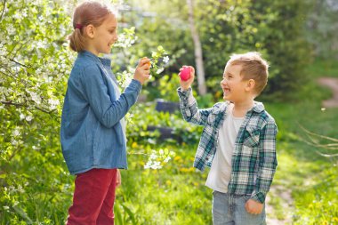Çocuklar bahar bahçesinde Paskalya yumurtası avında. Küçük çocuk ve anaokulu kızı açık havada oynuyorlar. Süslü yumurtaları kırıyorlar..