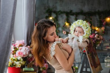 Bir anne, küçük kızını kollarında başında bir çelenkle tutar. Çiçekten şapkası olan güzel bir kadın ve çocuğun portresi. Bahar dekorasyonları.