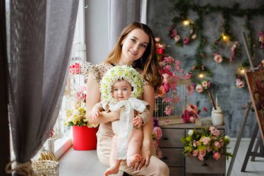 Bir anne, küçük kızını kollarında başında bir çelenkle tutar. Çiçekten şapkası olan güzel bir kadın ve çocuğun portresi. Bahar dekorasyonları.