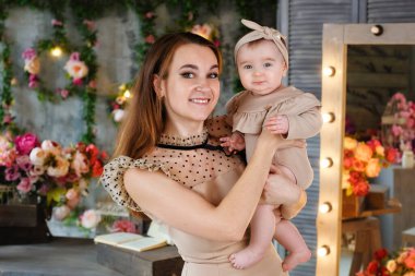 Bej elbiseli güzel, narin bir anne, küçük kızını stüdyoda, çiçekli bir vazonun arka planında kollarında tutuyor. Bahar süslemesinde bir kadın ve bir bebek.