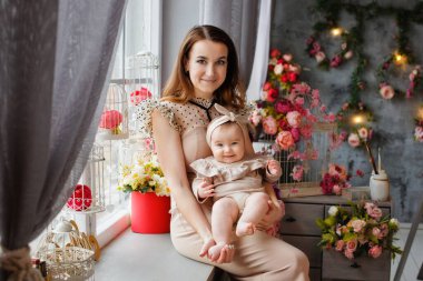 Bej elbiseli güzel, narin bir anne, küçük kızını stüdyoda, çiçekli bir vazonun arka planında kollarında tutuyor. Bahar süslemesinde bir kadın ve bir bebek.