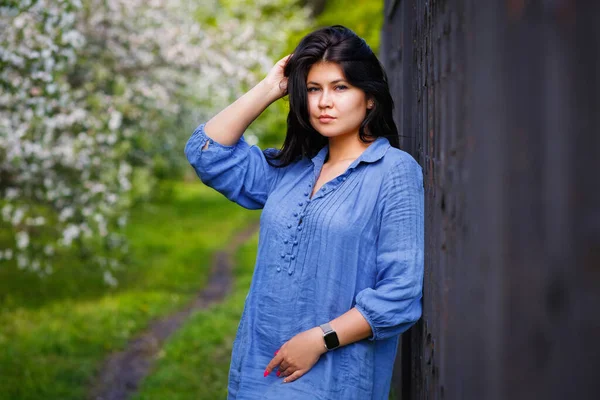 A young attractive woman plus size with dark hair in a blue dress poses in a spring blooming garden. Spring time.