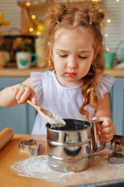Küçük komik bir bebek yardımcısı elinde bir testle oynuyor. Bebek yoğurmayı öğreniyor. Yetişkin anneye mutfakta yardım ediyor. Mutlu küçük kız ve ebeveyn annesi evde birlikte kurabiye yaparken eğleniyor.