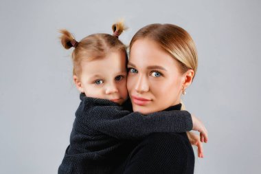Bir anne, kızını, stüdyoda gri bir arka planda sevgiyle kucaklıyor. Arka planda izole edilmiş koyu renk elbiseli bir kadın ve bir çocuk var..