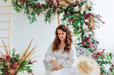 Beyaz uzun elbiseli güzel bir esmer, bahar çiçeklerinden yapılmış beyaz ışıklı bir odada bir sandalyede oturuyor..