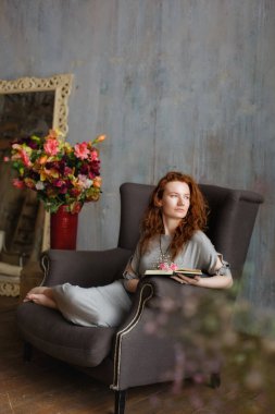 Çekici kızıl saçlı genç bir kadının portresi rahat gri bir sandalyede kitap okuyor. Yakınlarda bir vazo çiçek var..
