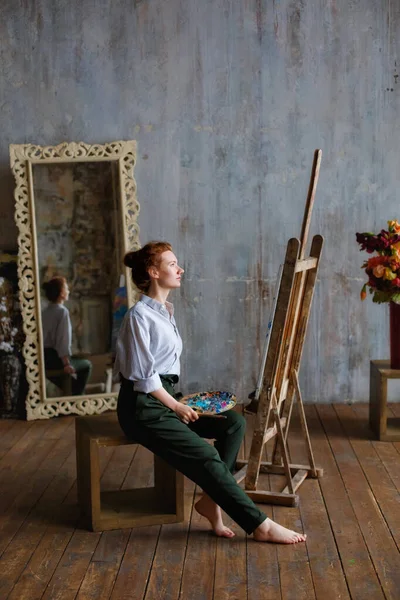 Portrait of a professional artist with a brush and palette, writing on canvas in the studio. A female artist draws in the workplace.