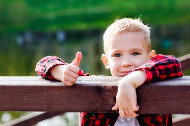 Tahta korkulukların yanındaki parkta bir çocuğun portresi. Kırmızı kareli tişörtlü bir çocuk baş parmak gösteriyor..