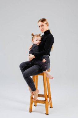 Bir anne, kızını, stüdyoda gri bir arka planda sevgiyle kucaklıyor. Arka planda izole edilmiş koyu renk elbiseli bir kadın ve bir çocuk var..