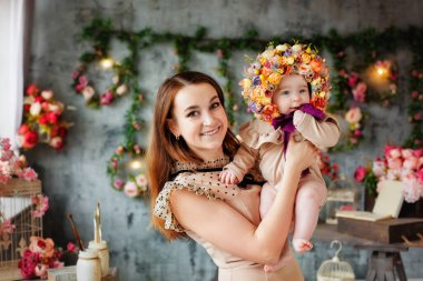 Bir anne, küçük kızını kollarında başında bir çelenkle tutar. Çiçekten şapkası olan güzel bir kadın ve çocuğun portresi. Bahar dekorasyonları.