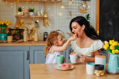 Parlak bir mutfağın arka planında neşeli bir aile. Anne ve kızı renkli çöreklerle eğleniyorlar. Bir kız bir kadını pastayla besler..