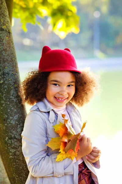 在一个阳光灿烂的日子里 一个戴着红帽子的非洲裔美国小女孩站在湖边的画像 一个女孩站在一棵树旁 手里拿着一束枫叶 — 图库照片