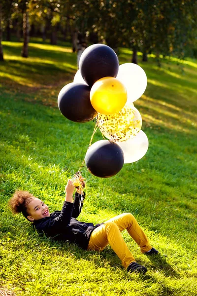 一个非洲小女孩正在一个室外公园里玩耍 他躺在草地上 手里拿着五彩缤纷的金 银气球 — 图库照片