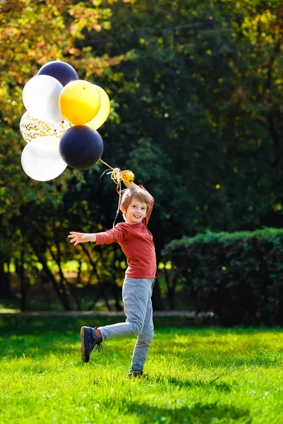 一个小学生 在秋天阳光灿烂的公园里拿着气球 一个红头发的男孩 长着一堆五颜六色的金球 黑球和银球 — 图库照片