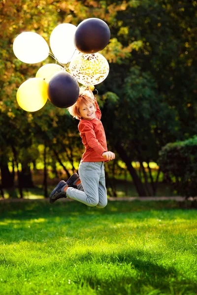 在一个阳光明媚的秋天公园里 一个小学生拿着气球跑来跑去 一个红头发的男孩 长着一堆五颜六色的金球 黑球和银球 — 图库照片