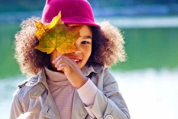 一个非洲裔美国女孩在一个公园里的肖像 在湖畔有一束秋天的叶子 她用枫叶闭上了一只眼睛 — 图库照片