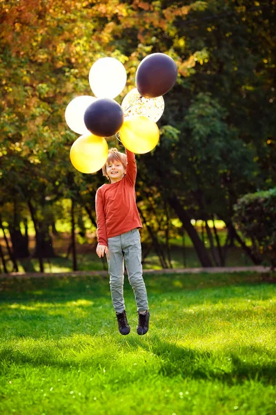 在一个阳光明媚的秋天公园里 一个小学生拿着气球跑来跑去 一个红头发的男孩 长着一堆五颜六色的金球 黑球和银球 — 图库照片