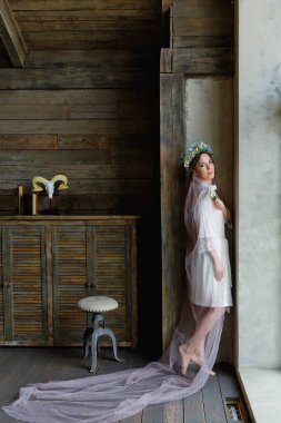 Düğün saç stili ve makyajlı, duvağı ve penceresinde çiçek çelengi olan güzel bir gelinin portresi. Gelinin sabahı.