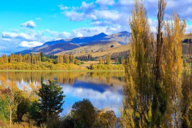 Hayes Gölü, Otago Bölgesi, Güney Adası, Yeni Zelanda. Gölün sakin suları çevredeki dağları ve sonbahar ağaçlarını yansıtır.
