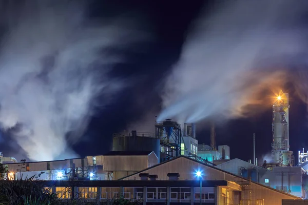 A pulp and paper factory at night, with steam pouring from the chimneys