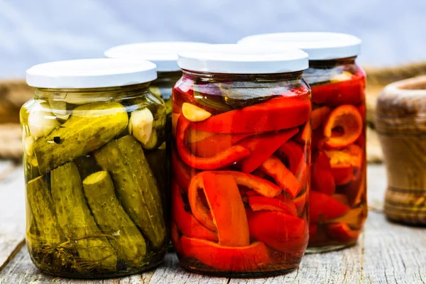 Glass jars with pickled red bell peppers and pickled cucumbers (pickles) isolated. Jars with variety of pickled vegetables. Preserved food concept in a rustic composition.