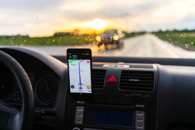 Araba gösterge panelindeki akıllı telefondan dalga haritası uygulamasını kullanan sürücü yağmurlu bir akşamda gün batımında şehir trafiğinde doğru yolu göstermek için harita uygulaması kullanıyor. Bükreş, Romanya, 2020.