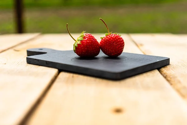 stock image Close up of two strawberries on small black cutting board isolated outdoor on wooden table.