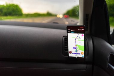 Araba gösterge panelindeki akıllı telefondan dalga haritası uygulamasını kullanan sürücü yağmurlu bir akşamda gün batımında şehir trafiğinde doğru yolu göstermek için harita uygulaması kullanıyor. Bükreş, Romanya, 2020.