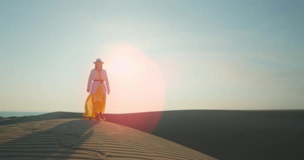 Χαλαρή γυναίκα που περπατάει στην έρημο το ηλιοβασίλεμα. Κορίτσι με καπέλο και κίτρινο φόρεμα — Αρχείο Βίντεο