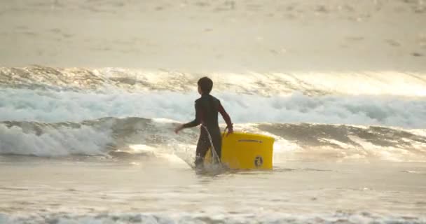 Ноябрь 2020 г. Молодой спортсмен бежит к океану с желтой доской для серфинга — стоковое видео