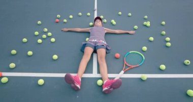 Tenis kortunda yatan kız. Toplarla ve küreklerle çevrili.