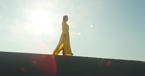 Wanita bergaya dengan gaun kuning berjalan di atas bukit pasir dalam gerakan lambat — Stok Video