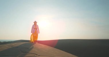 Gün batımında çölde yürüyen rahat bir kadın. Şapkalı ve sarı elbiseli kız.
